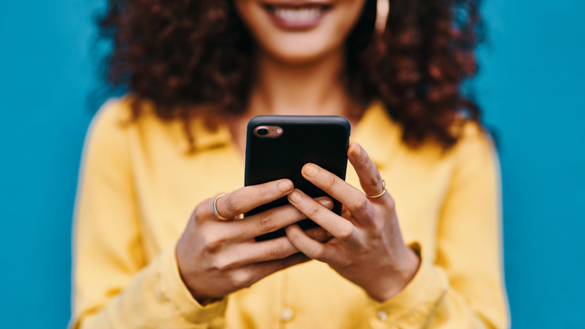 A person with curly hair wearing a yellow shirt smiles while holding and looking at a smartphone against a blue background.